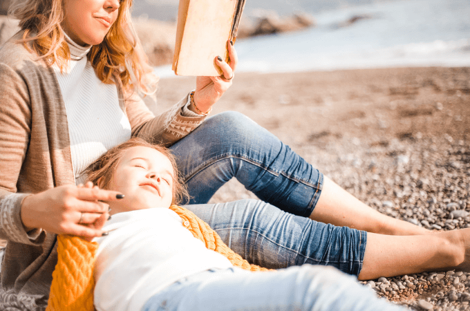 image of little girl on the beach with her mother and mom is reading to the girl from a book - suggesting