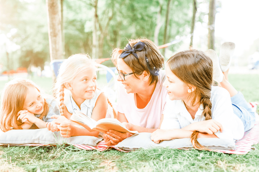Happy mother and three pretty daughters reading outside in the sunshine - concept of read aloud books or books for summer