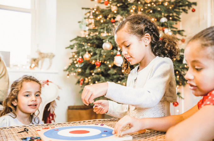 Photo of three young girls playing a table top game with a Christmas tree in the background - suggestive of fun family Christmas games