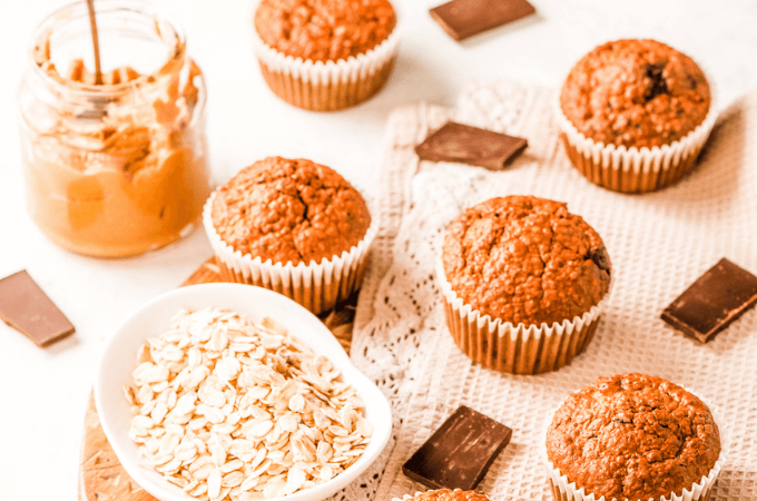 An image of peanut butter muffins with chunks of chocolate, a jar of oats, and a jar of peanut butter suggesting delicious homemade peanut butter muffins